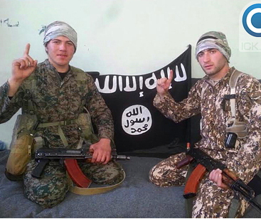 Two teenage boys seated, dressed in military fatigues with machine guns, IS flag in background